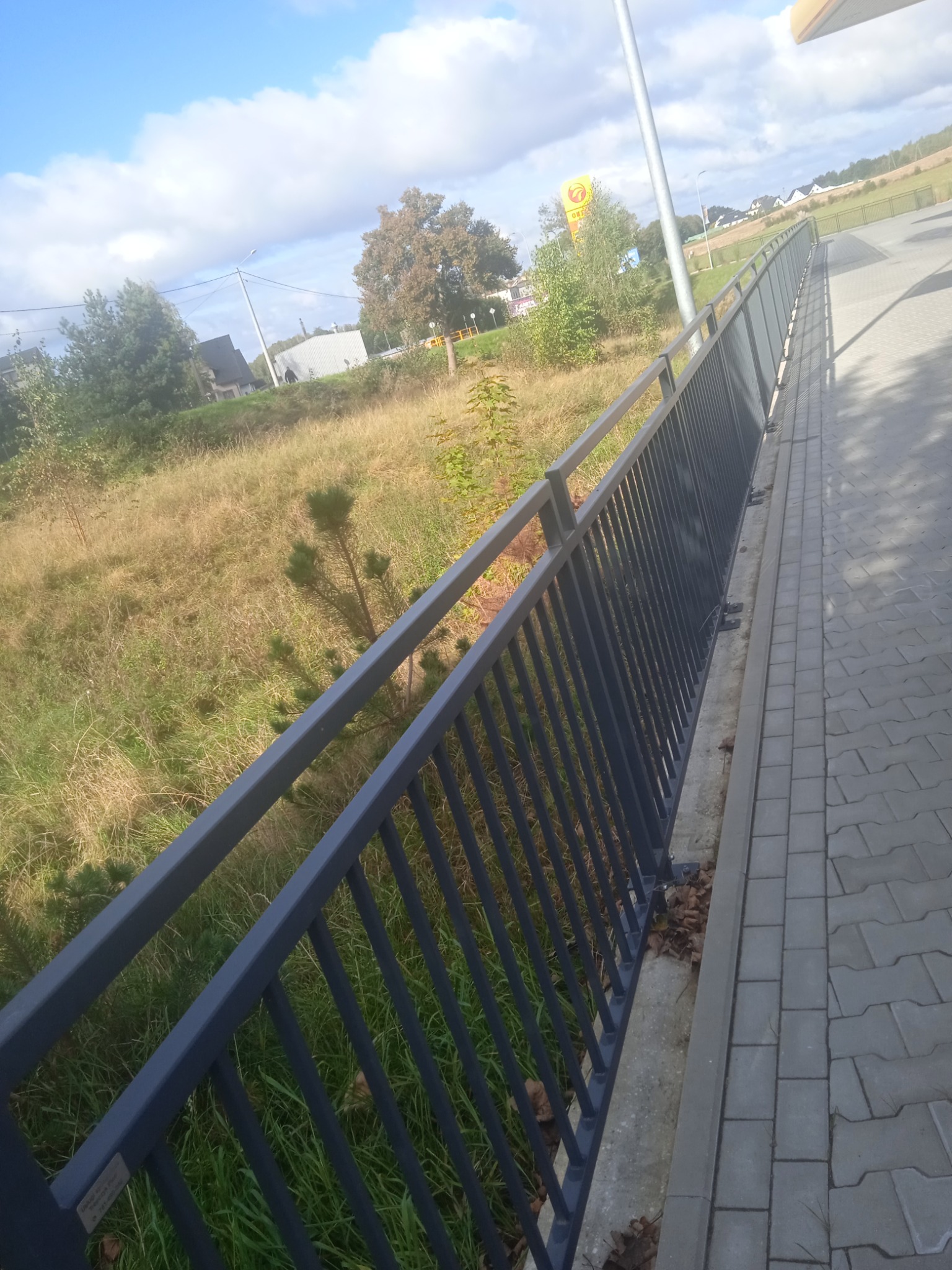 Długa, szara barierka z metalowych prętów, zamontowana wzdłuż chodnika. W tle trawa i drzewa pod błękitnym niebem. Widok z perspektywy lekko z góry.