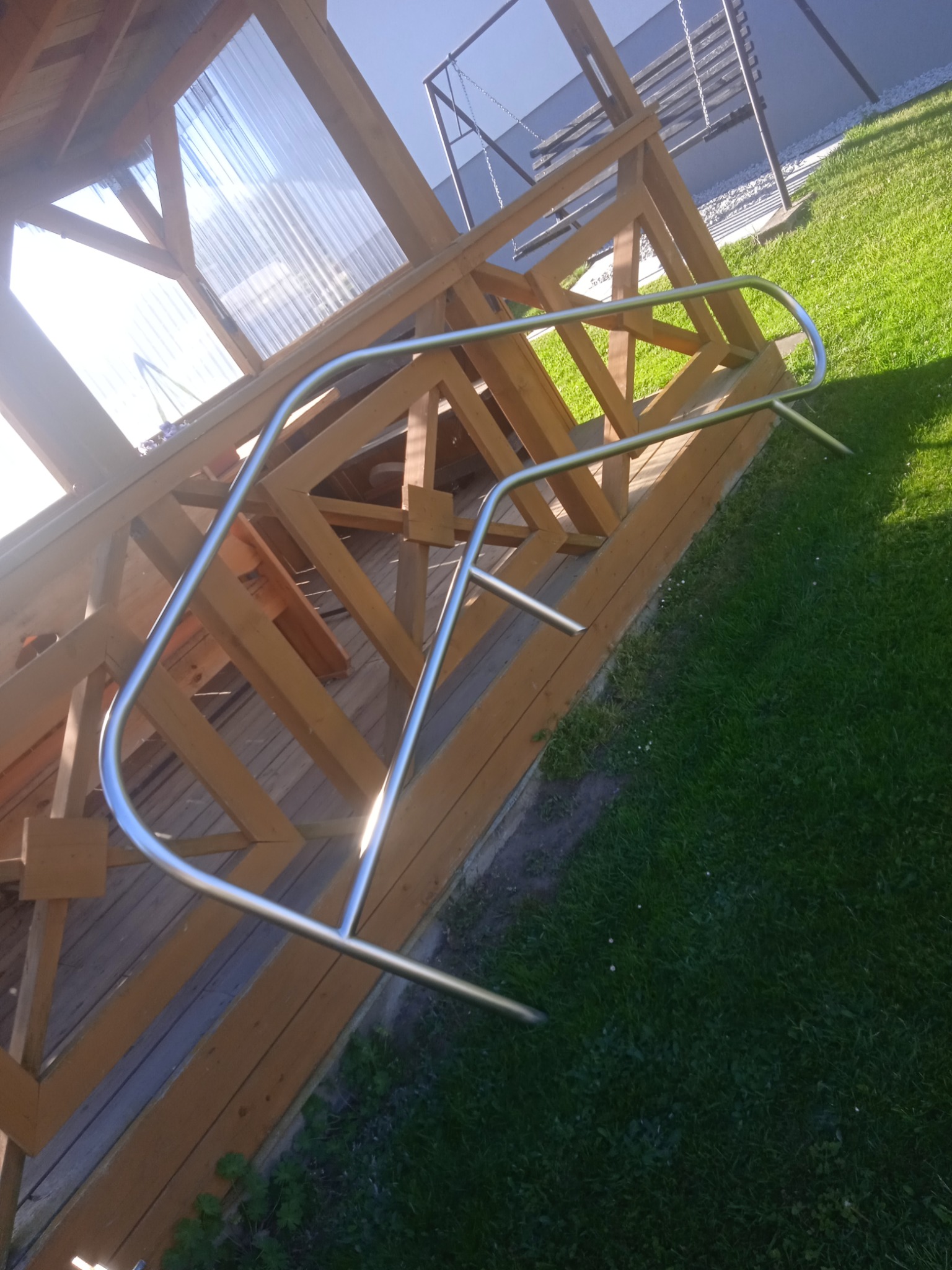 Metalowa poręcz przy drewnianej konstrukcji zadaszenia, widok z boku, trawa w tle, słoneczny dzień, lekki cień. Poręcz zamontowana na schodach.