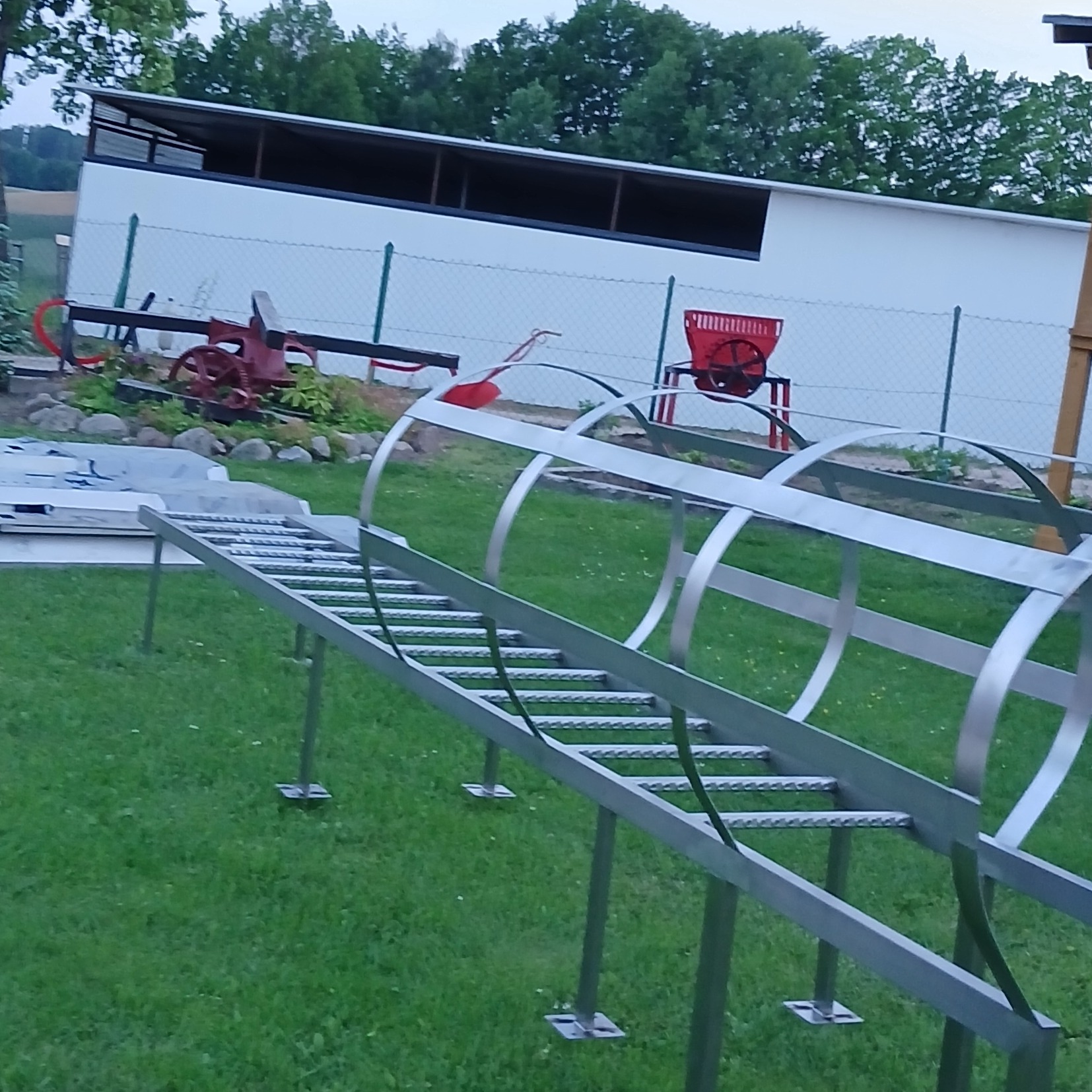 Metalowa konstrukcja przypominająca zadaszony mostek na trawniku, wykonana z profili i płaskowników, z widocznymi elementami mocującymi do podłoża.