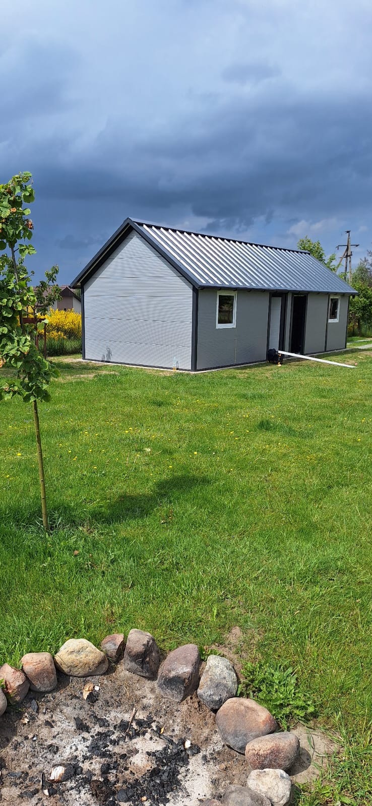 Szary garaż blaszany z metalowym dachem na zielonej trawie, widok z przodu, pochmurne niebo w tle. Ognisko z kamieni na pierwszym planie.