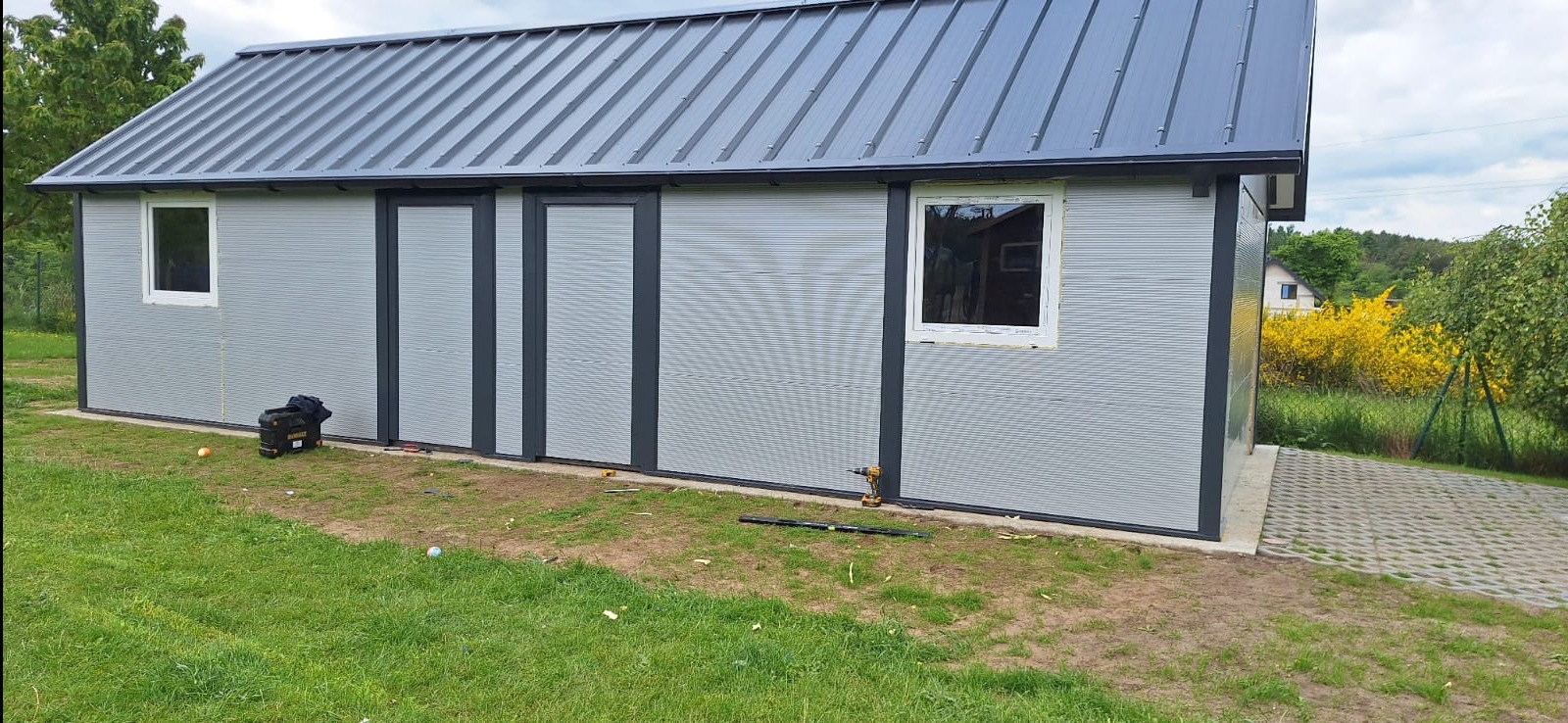 Szary garaż blaszany z metalowym dachem, widoczne narzędzia i skrzynka narzędziowa na trawie. Ukończona konstrukcja z zamontowanymi drzwiami i oknami.