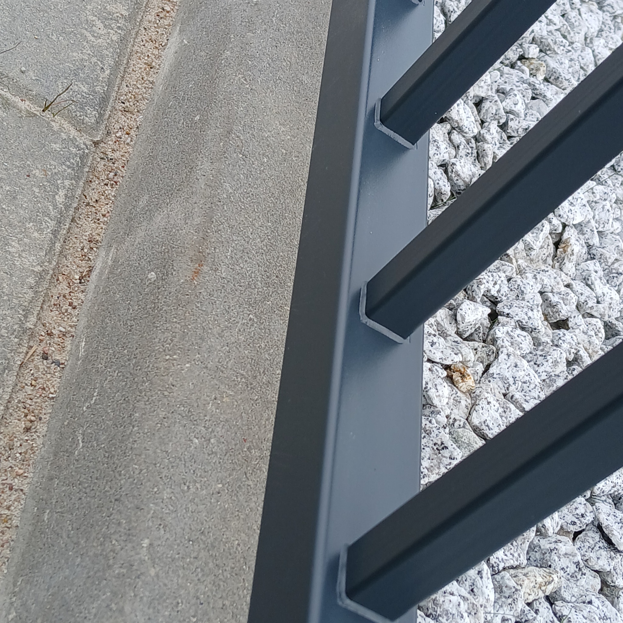 Fragment nowoczesnej, grafitowej balustrady z kwadratowych profili, zamontowanej na tle białych kamieni i betonowej kostki. Detal spawu widoczny u podstawy słupka.