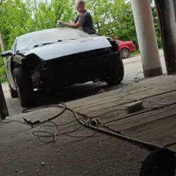 Przygotowanie karoserii samochodu do lakierowania w warsztacie, widoczne przewody i narzędzia pneumatyczne na posadzce, w tle inny samochód.