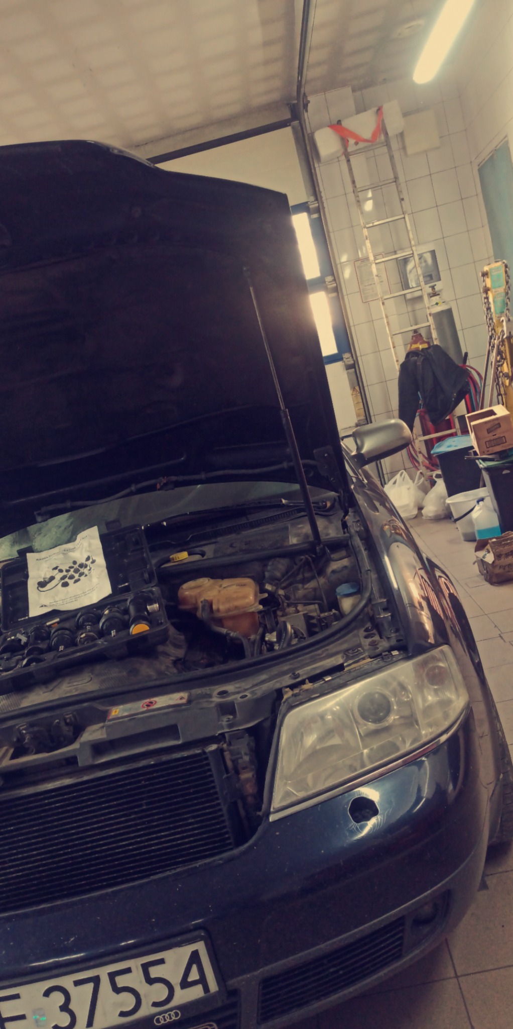 Otwarta maska samochodu Audi w warsztacie, widoczny silnik z zestawem narzędzi diagnostycznych na wierzchu, tło z drabiny i akcesoriów warsztatowych.