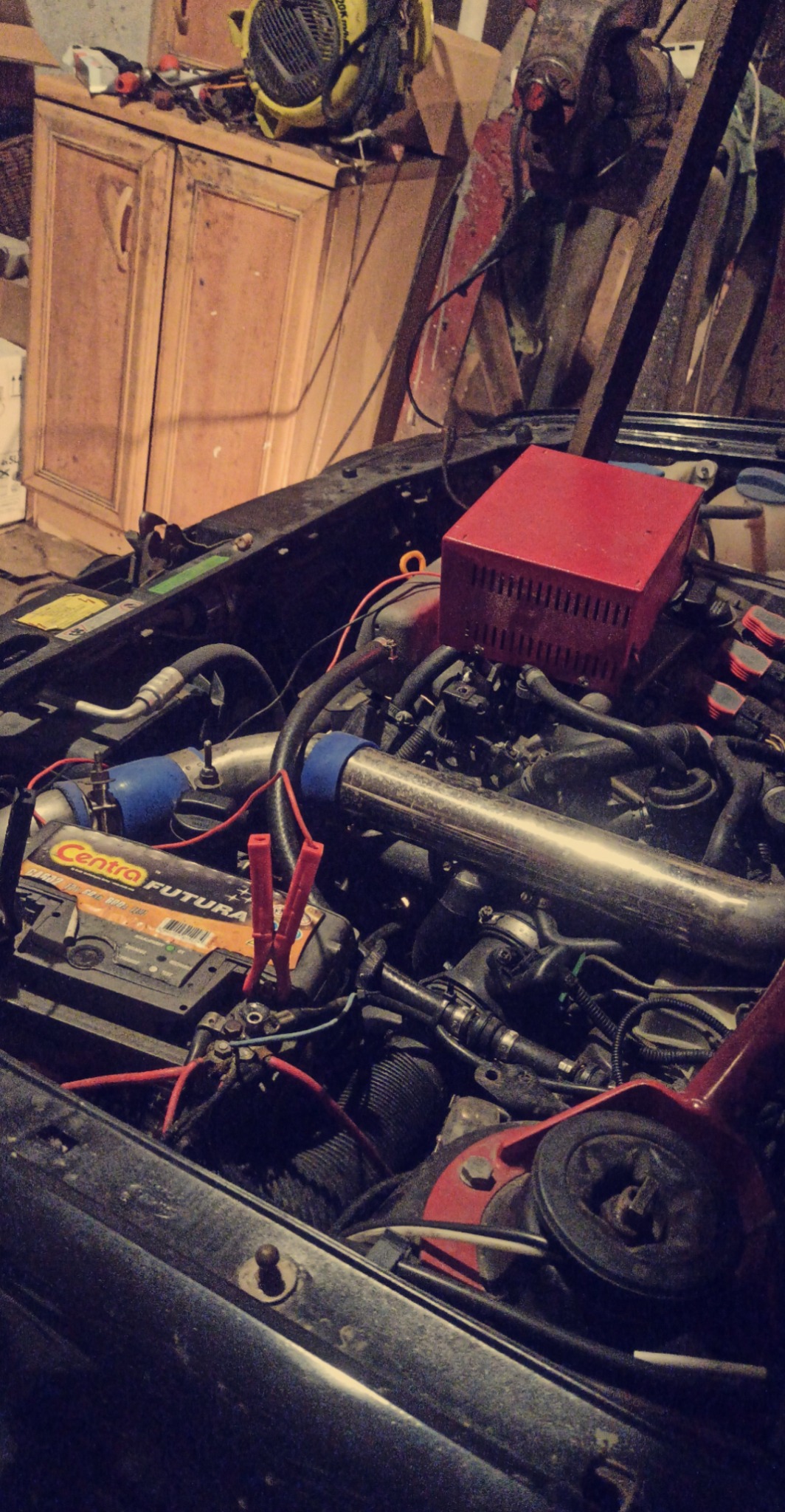 Silnik samochodowy z podłączonymi przewodami rozruchowymi, widoczna skrzynka elektryczna w kolorze czerwonym i fragment warsztatu w tle.