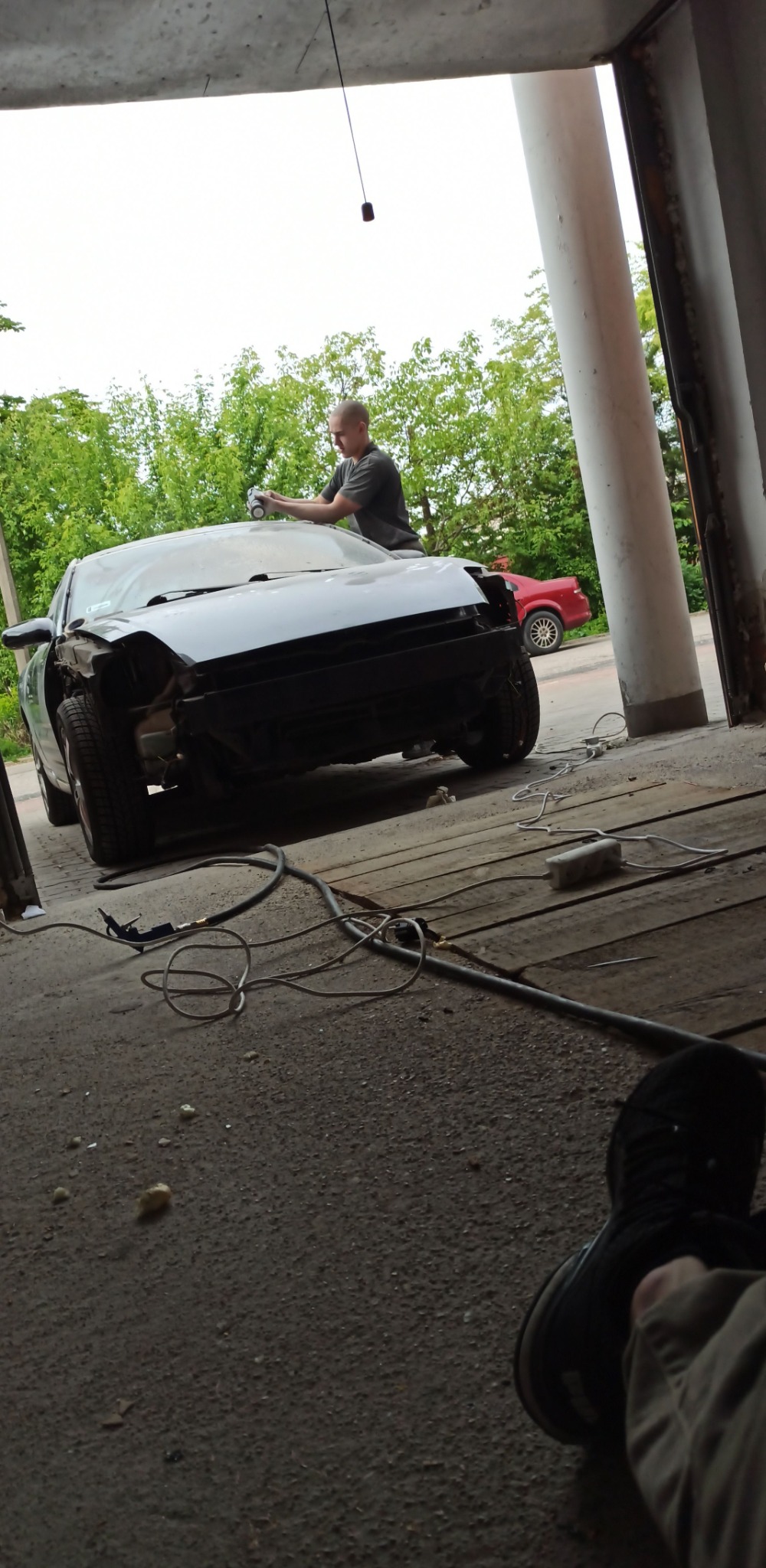 Przygotowanie karoserii samochodu do lakierowania w warsztacie, widoczne przewody i narzędzia pneumatyczne na posadzce, w tle inny samochód.