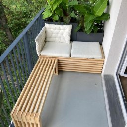 Meblitom - Drewniana ławka i siedzisko z poduszkami na balkonie, obok donica z zielonymi roślinami.