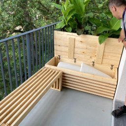 Meblitom - Drewniana skrzynia balkonowa z roślinami i ławką z listew, otwierana przez mężczyznę na balkonie z szarą podłogą i metalową balustradą.