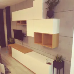 Nowoczesna zabudowa ściany w salonie z białymi szafkami i drewnianymi akcentami, wiszący telewizor oraz rośliny doniczkowe w czarnych osłonkach.