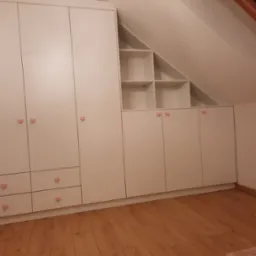 Biała zabudowa meblowa pod skosem dachu, z szafkami i otwartymi półkami, ozdobiona uchwytami w kształcie różowych serduszek, na drewnianej podłodze.