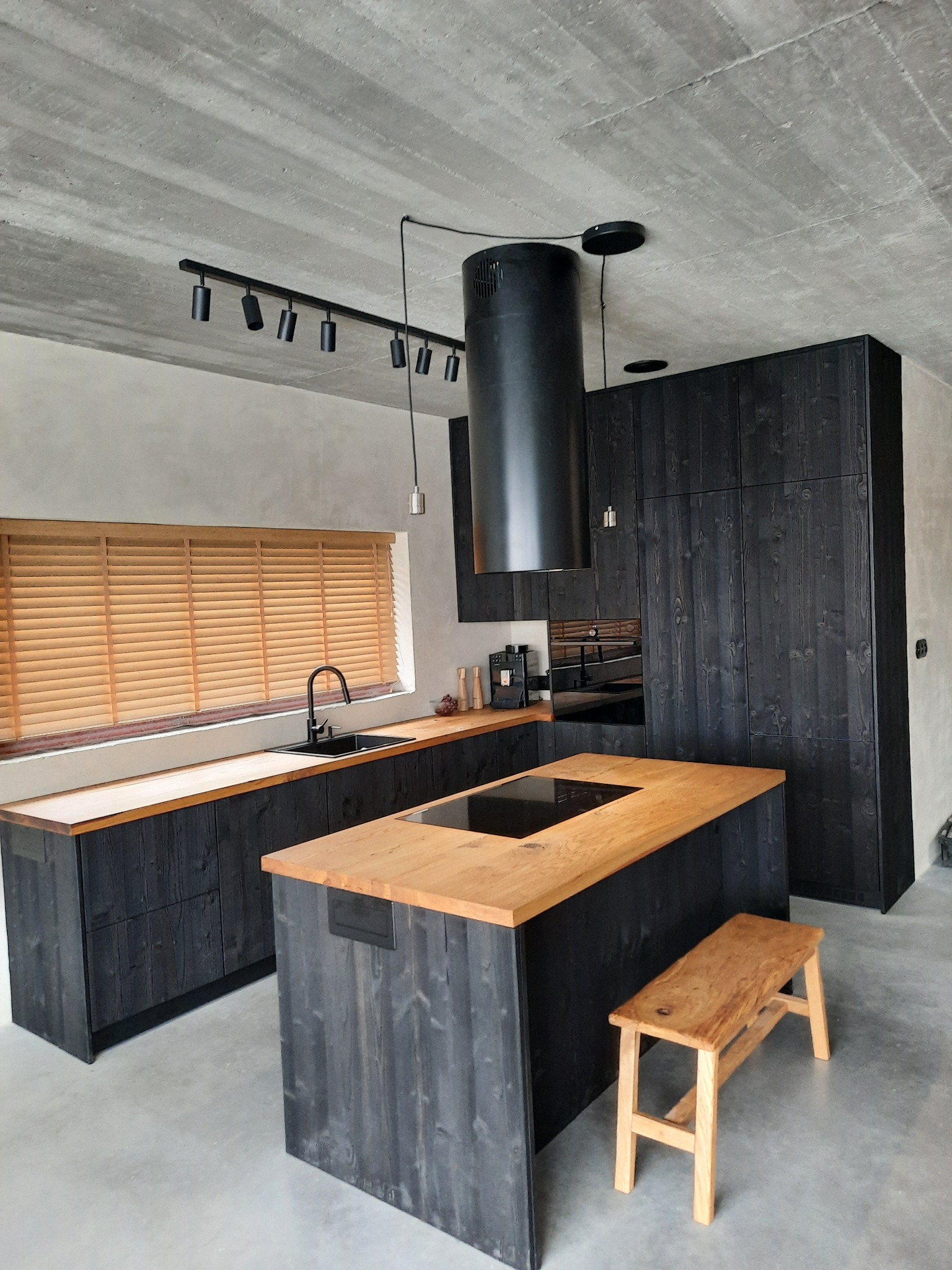 Nowoczesna kuchnia z czarnymi szafkami i drewnianymi blatami, surowy beton na suficie, wyspa kuchenna z drewnianym blatem i drewniany stołek.
