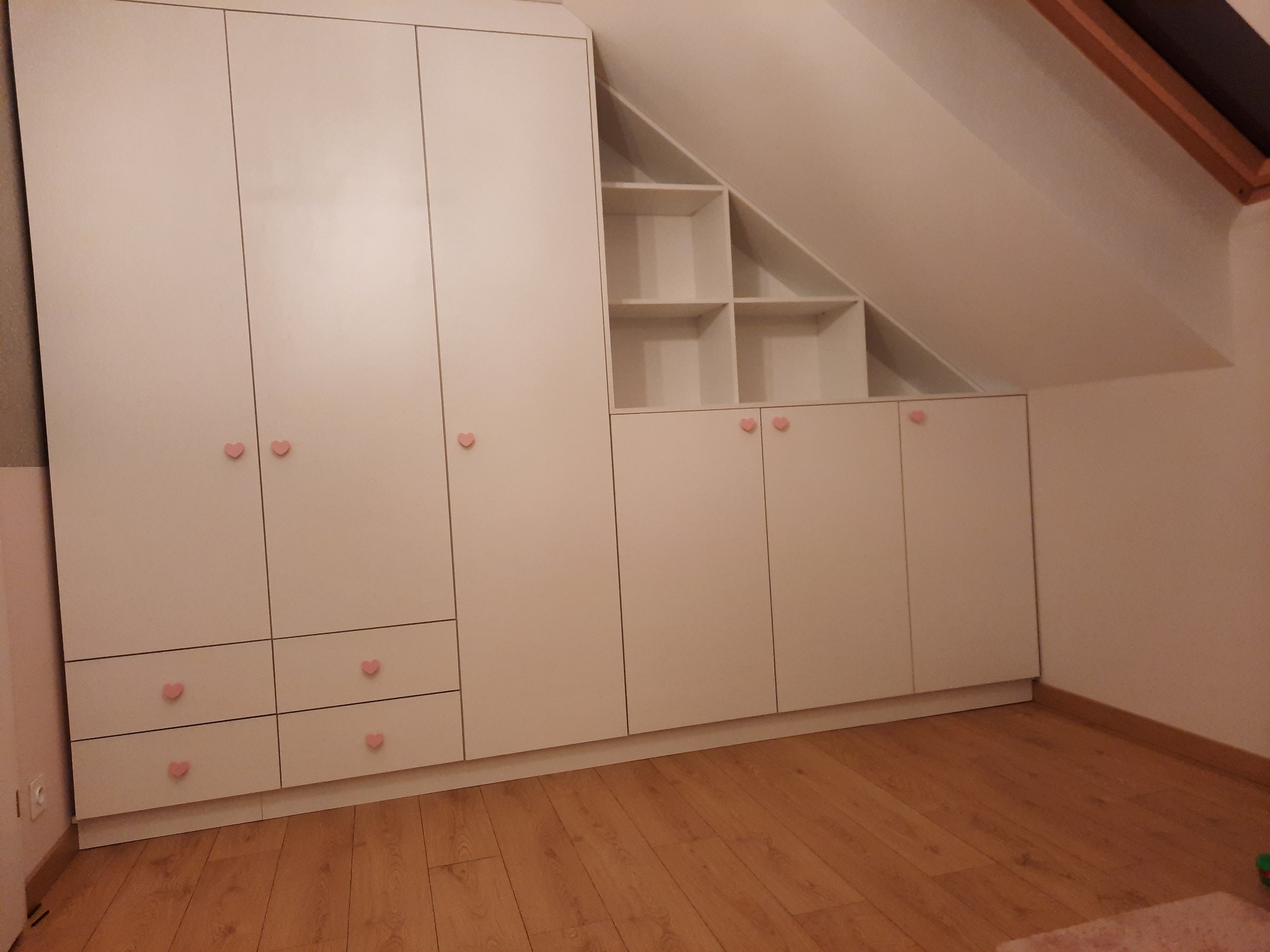 Biała zabudowa meblowa pod skosem dachu, z szafkami i otwartymi półkami, ozdobiona uchwytami w kształcie różowych serduszek, na drewnianej podłodze.