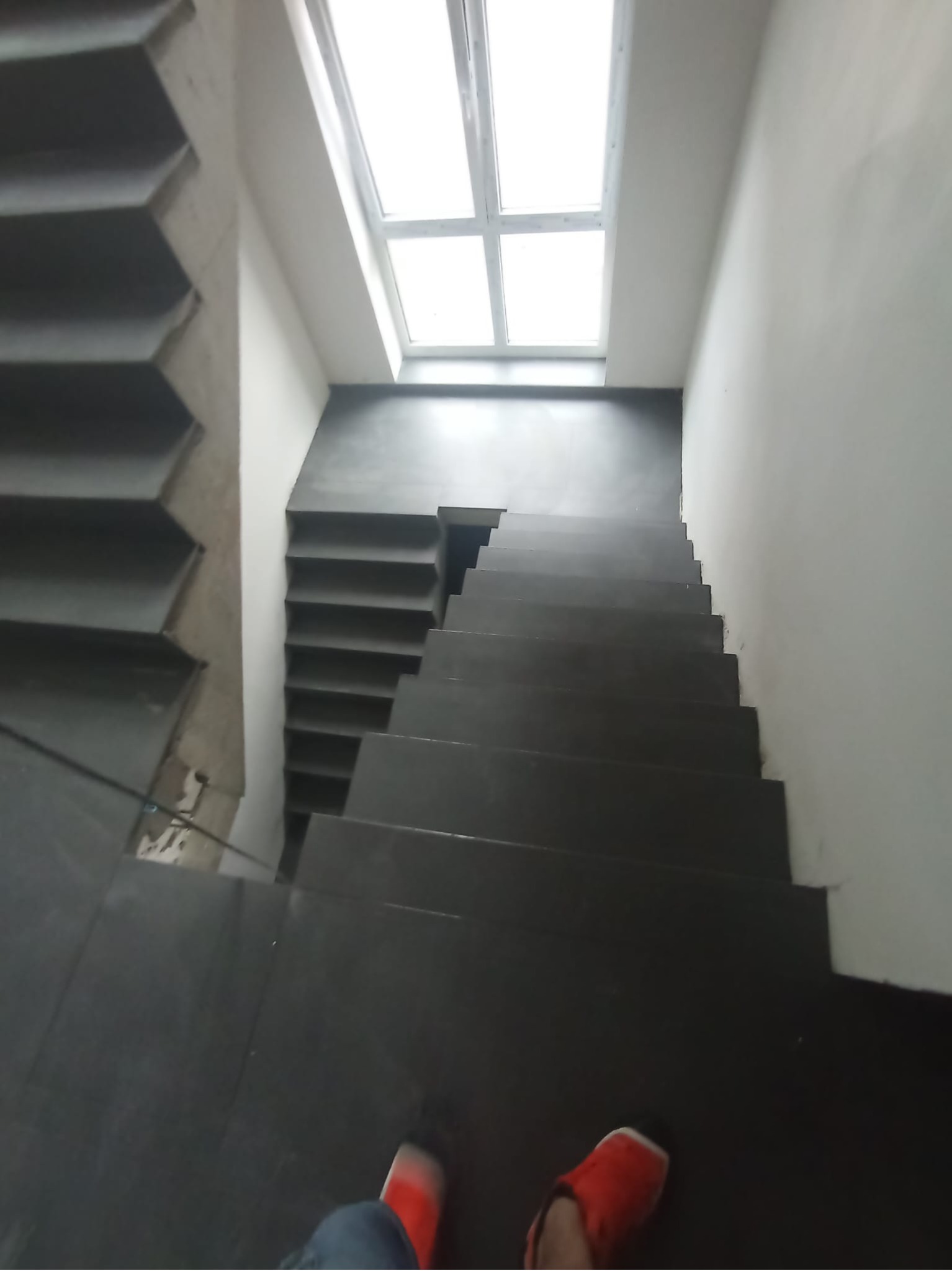 Ciemne, wykończone schody z jasnym oknem dachowym na szczycie, ujęcie z góry. Po lewej widoczna surowa betonowa konstrukcja, po prawej biała ściana.