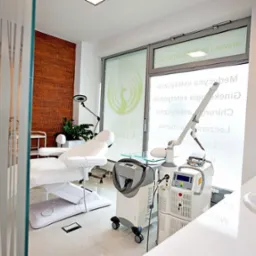 Jasne wnętrze gabinetu z kozetką, laserem zabiegowym Zimer i umywalką, widoczne logo Revival Clinic na oknie.