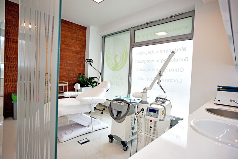 Jasne wnętrze gabinetu z kozetką, laserem zabiegowym Zimer i umywalką, widoczne logo Revival Clinic na oknie.