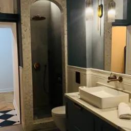 Nowoczesna łazienka z kabiną prysznicową wykończoną mozaiką, umywalką nablatową i szafką w kolorze granatowym z złotymi uchwytami, oświetlona złotymi kinkietami.