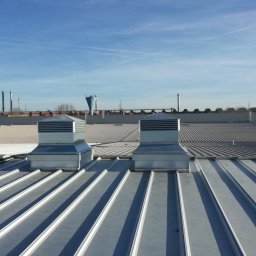 Mat Max Instalacje. Mateusz Szeliga - Dwa metalowe wywietrzniki dachowe na przemysłowym dachu z blachy trapezowej, w tle kominy fabryczne i błękitne niebo z smugami kondensacyjnymi.