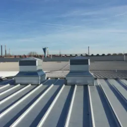 Dwa metalowe wywietrzniki dachowe na przemysłowym dachu z blachy trapezowej, w tle kominy fabryczne i błękitne niebo z smugami kondensacyjnymi.