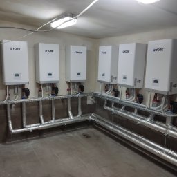Mat Max Instalacje. Mateusz Szeliga - Rząd białych, wiszących kotłów grzewczych YORK z podłączonymi rurami w izolacji, zamontowanych w pomieszczeniu z szarą podłogą i beżowymi ścianami.