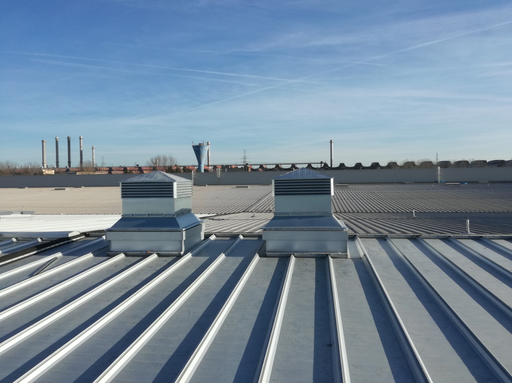 Dwa metalowe wywietrzniki dachowe na przemysłowym dachu z blachy trapezowej, w tle kominy fabryczne i błękitne niebo z smugami kondensacyjnymi.