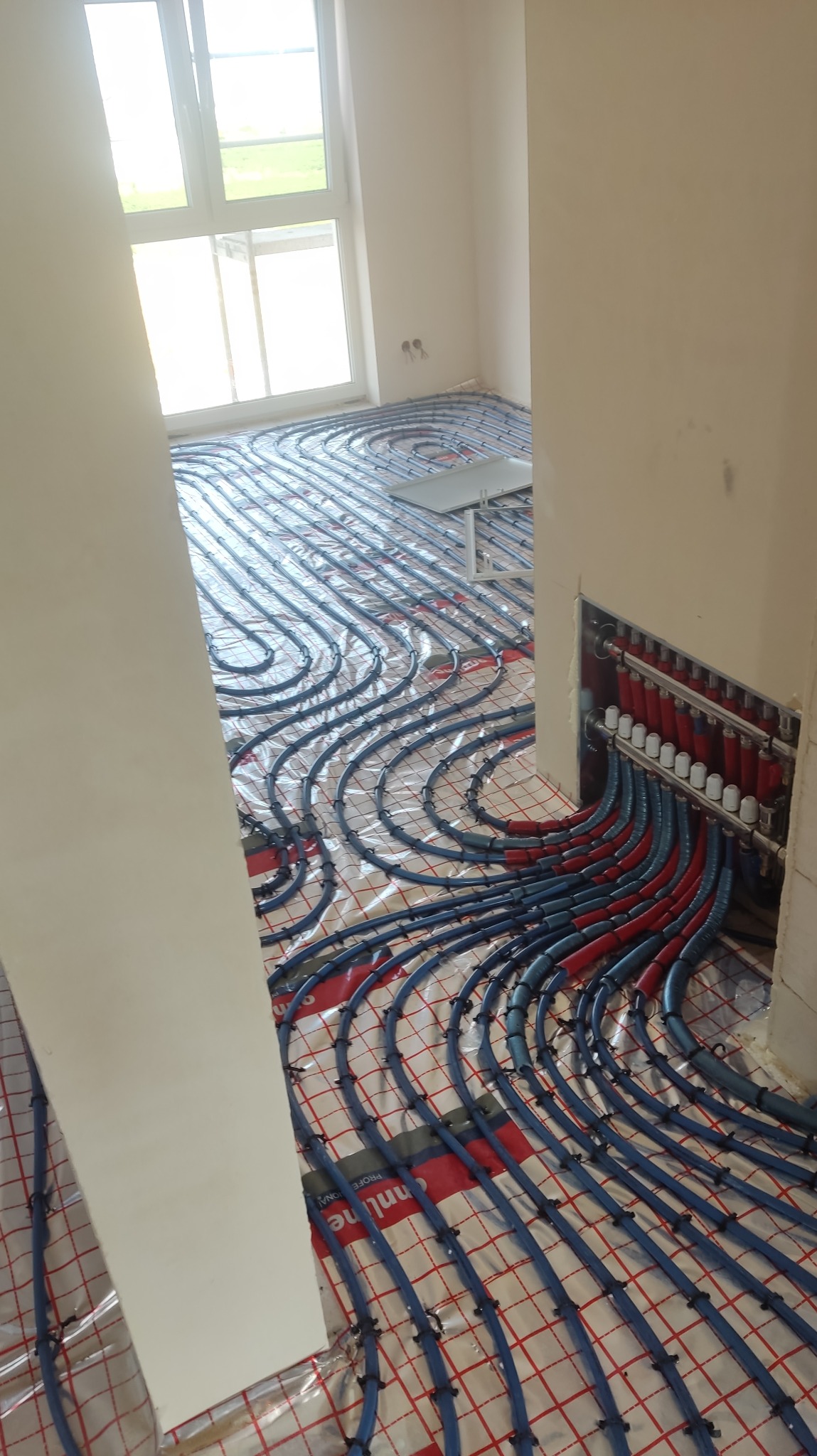 Instalacja ogrzewania podłogowego w nowym budynku: widoczne niebieskie rurki układane na folii z siatką, kolektor z czerwonymi i niebieskimi przewodami w ścianie, oraz fragment okna w tle.