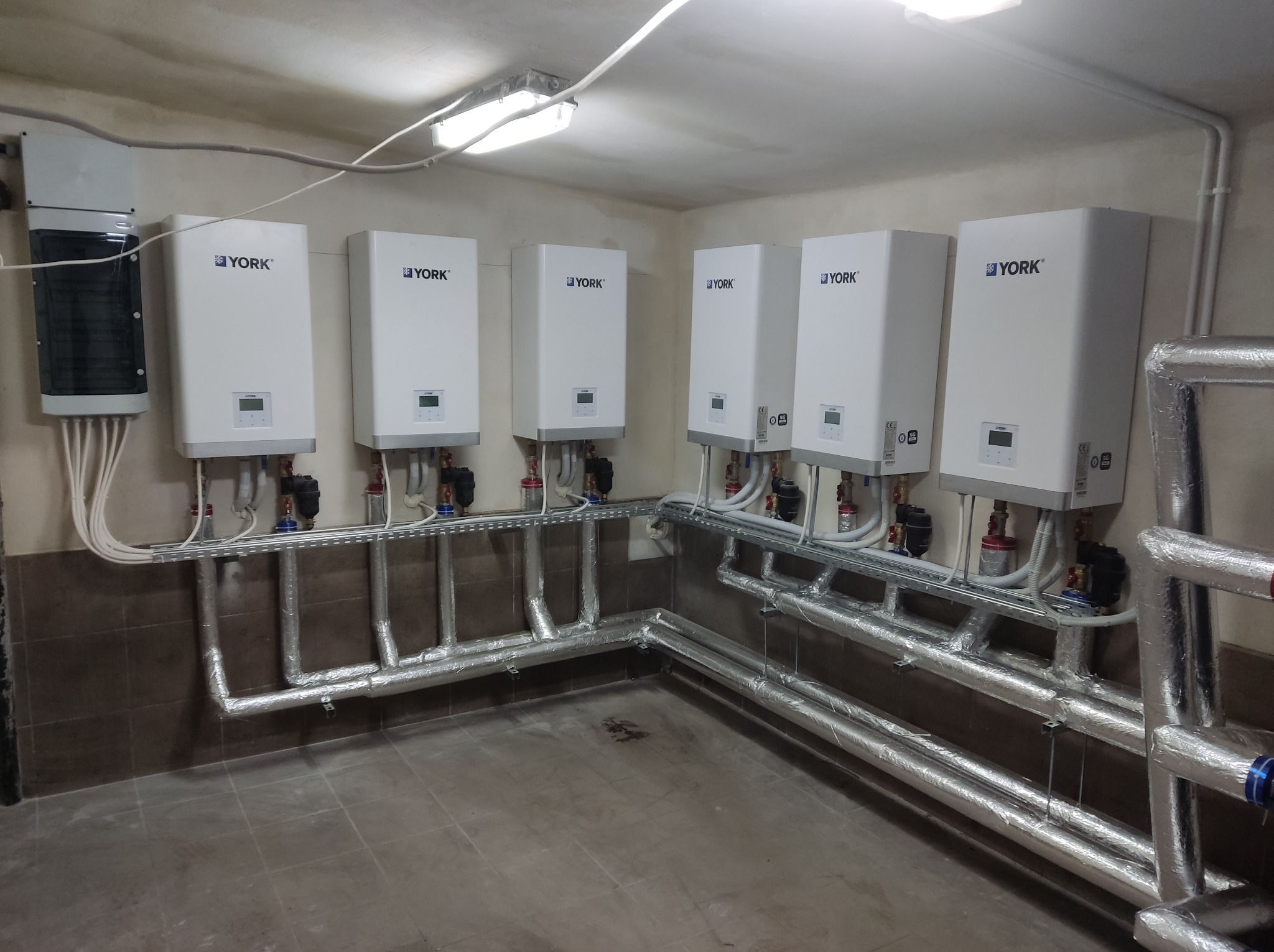 Rząd białych, wiszących kotłów grzewczych YORK z podłączonymi rurami w izolacji, zamontowanych w pomieszczeniu z szarą podłogą i beżowymi ścianami.