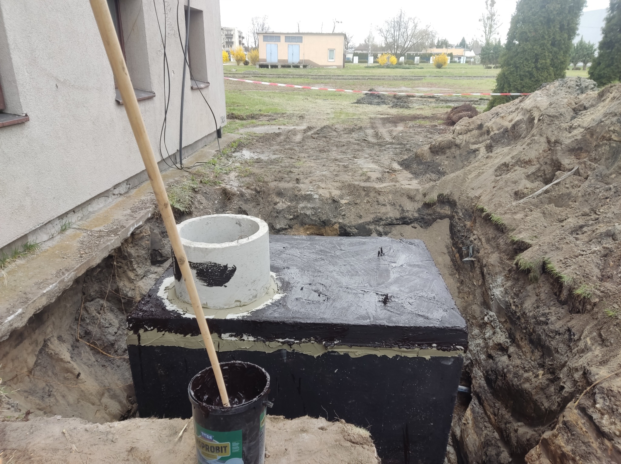 Instalacja betonowego zbiornika w wykopie, zabezpieczonego czarną izolacją, z widocznym betonowym kręgiem i wiadrem z substancją uszczelniającą.