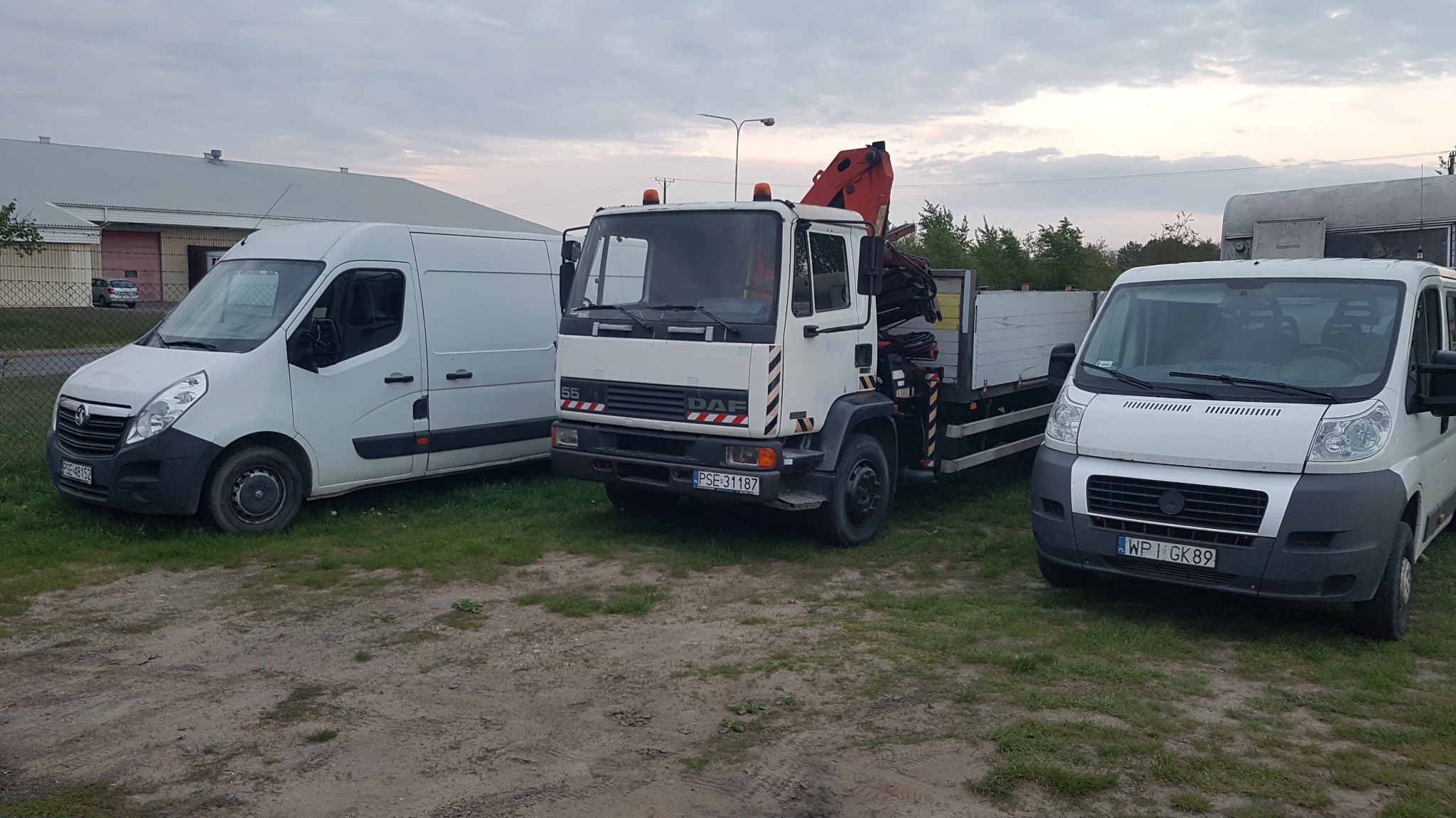 Trzy zaparkowane białe samochody dostawcze, w tym jeden ciężarowy z dźwigiem, stoją na ziemi z trawą przed budynkiem w pochmurny dzień.