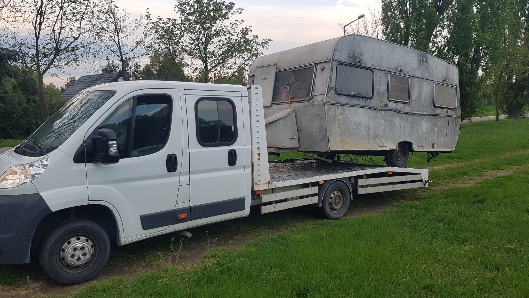 Biała laweta przewozi starą, srebrną przyczepę kempingową na tle zielonej trawy i drzew.