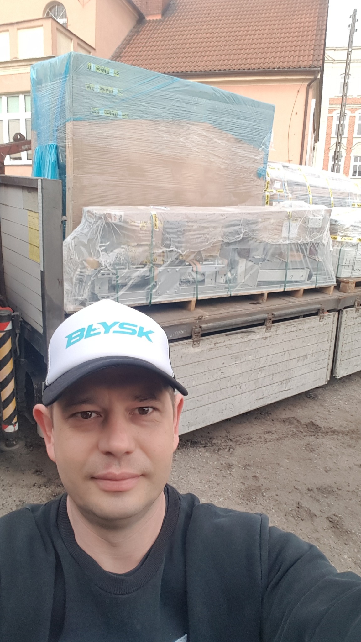 Autoportret mężczyzny w czapce z daszkiem na tle ciężarówki z załadowanym towarem na paletach, zabezpieczonym folią, budynki w tle.