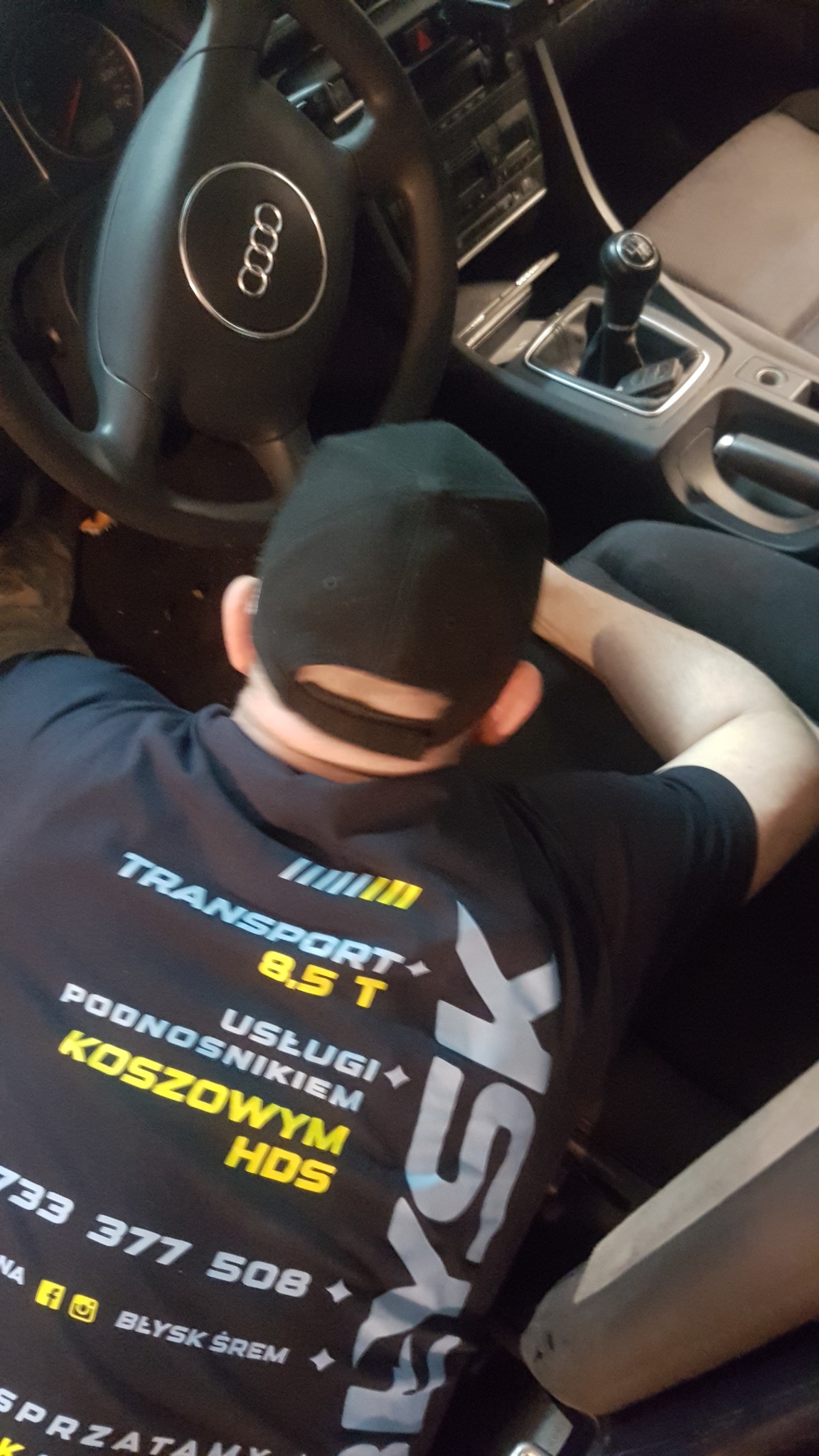 Mężczyzna w czarnej koszulce z napisem 'TRANSPORT 8.5 T' siedzi za kierownicą samochodu marki Audi, widoczna kierownica, deska rozdzielcza i dźwignia zmiany biegów.