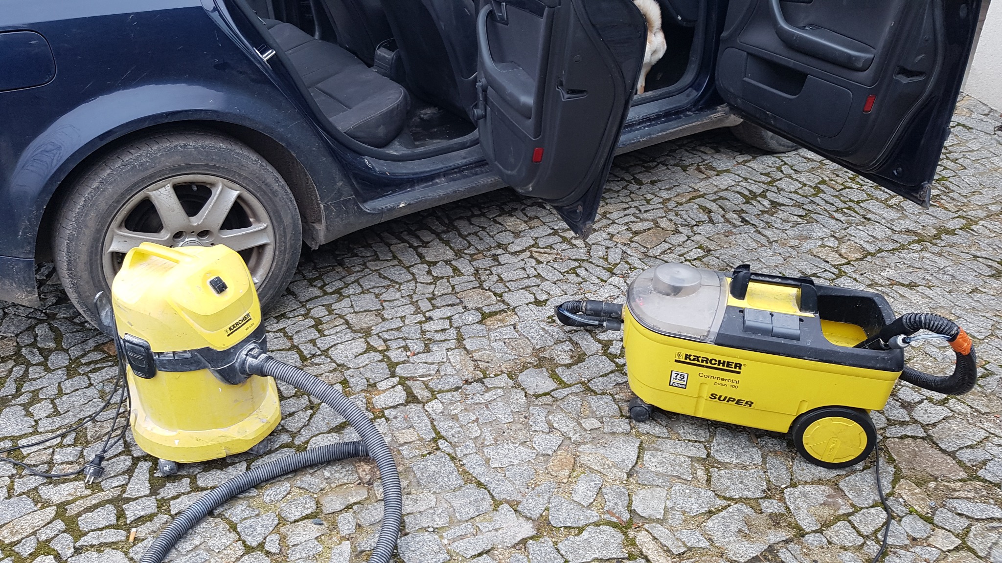 Dwa żółte odkurzacze Kärcher stoją na brukowanej nawierzchni obok otwartego, granatowego samochodu osobowego z widocznym fragmentem wnętrza.