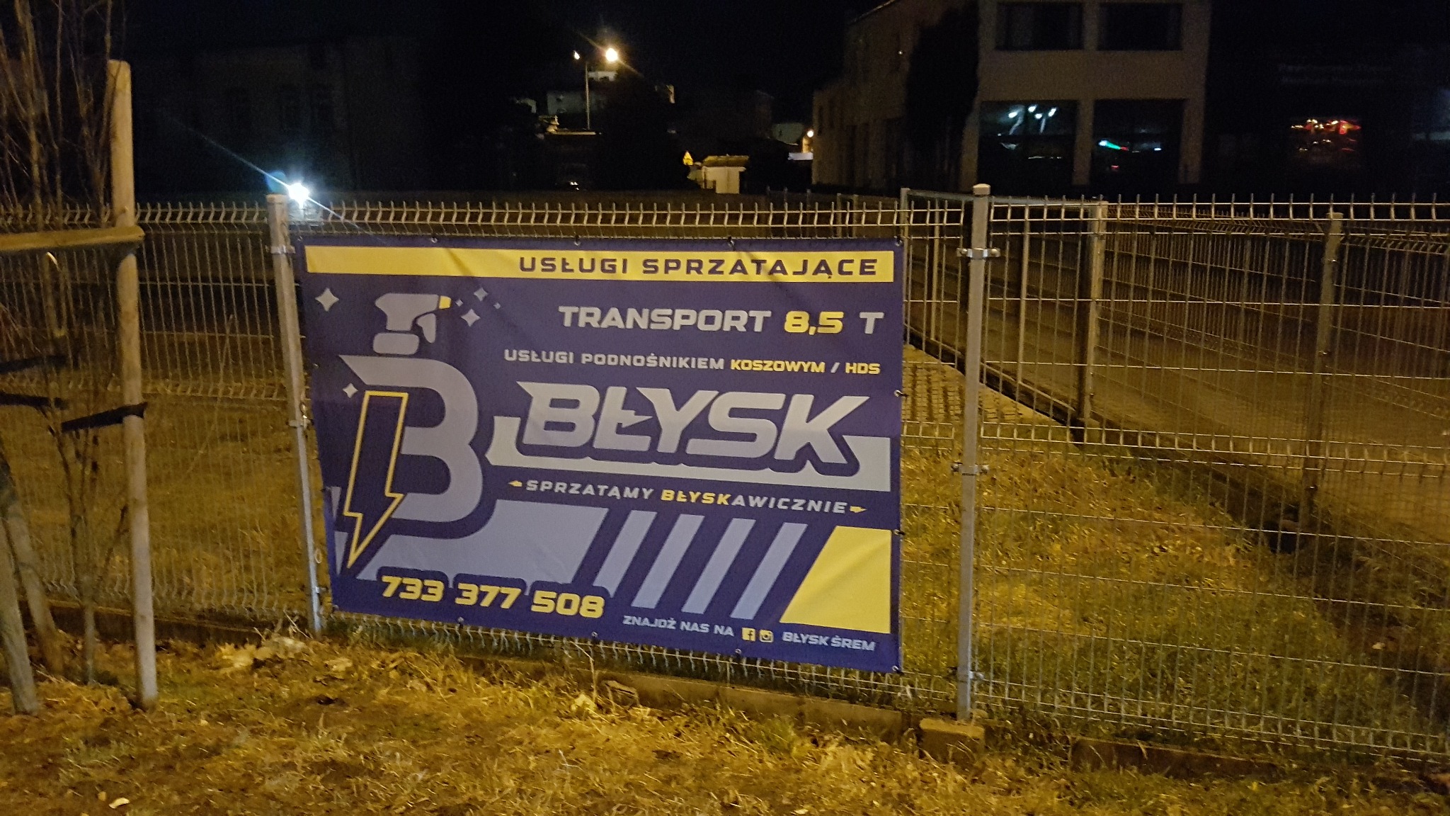 Baner reklamowy firmy 'Błysk' zawieszony na ogrodzeniu, oferującej usługi sprzątające, transport 8,5T oraz usługi podnośnikiem koszowym/HDS, widoczny numer telefonu i adres Facebook. Zdjęcie...