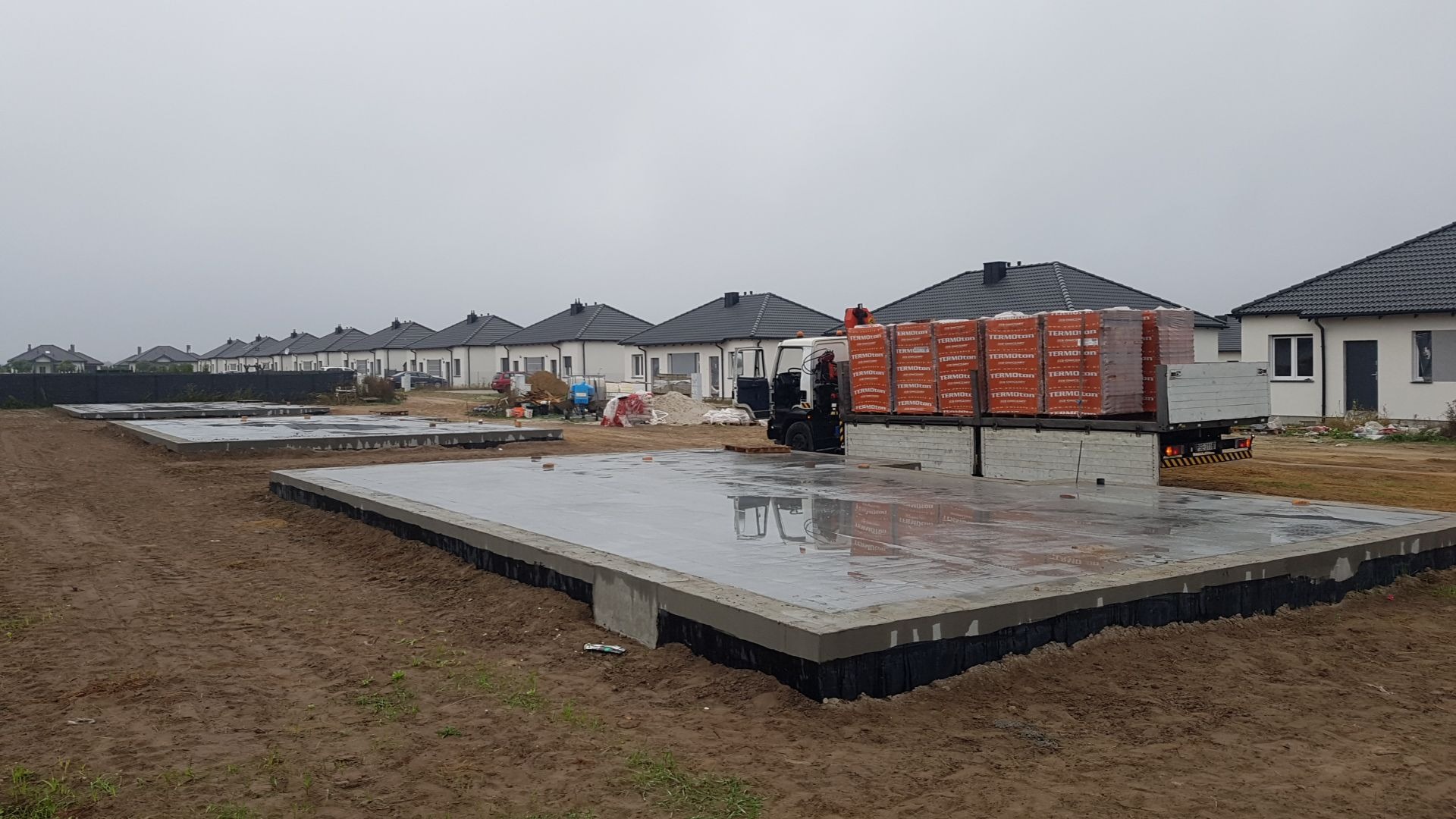 Przygotowane fundamenty pod budowę osiedla domów jednorodzinnych; na jednym z fundamentów zaparkowana ciężarówka z materiałami budowlanymi - cegłami Termoton.