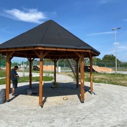 Przemysław Strach - Ośmioboczna konstrukcja z ciemnym, spadzistym dachem, prawdopodobnie w trakcie montażu, z pracownikiem w tle i elementami placu zabaw skateparku w oddali.