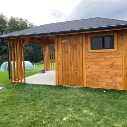 Przemysław Strach - Nowoczesna, drewniana konstrukcja z zadaszeniem, przypominająca mały domek lub pawilon ogrodowy, z wyraźnymi słojami drewna, czarnym dachem i rynnami, otoczona zielonym trawnikiem i drzewami w tle.