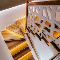 Przemysław Strach - Nowoczesne schody z podświetlanymi stopniami i ażurową, białą balustradą z drewnianym wykończeniem, widok z góry.