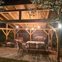 Drewniana konstrukcja z oświetleniem, zadaszona, z meblami ogrodowymi na kamiennej posadzce, otoczona drzewami, oświetlona lampami zewnętrznymi i girlandą żar&oacute;wek.
