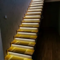 Nowoczesne schody z podświetleniem LED na każdym stopniu, widoczne z dołu do góry, z teksturą ściany po lewej stronie i ciemną podłogą po prawej.