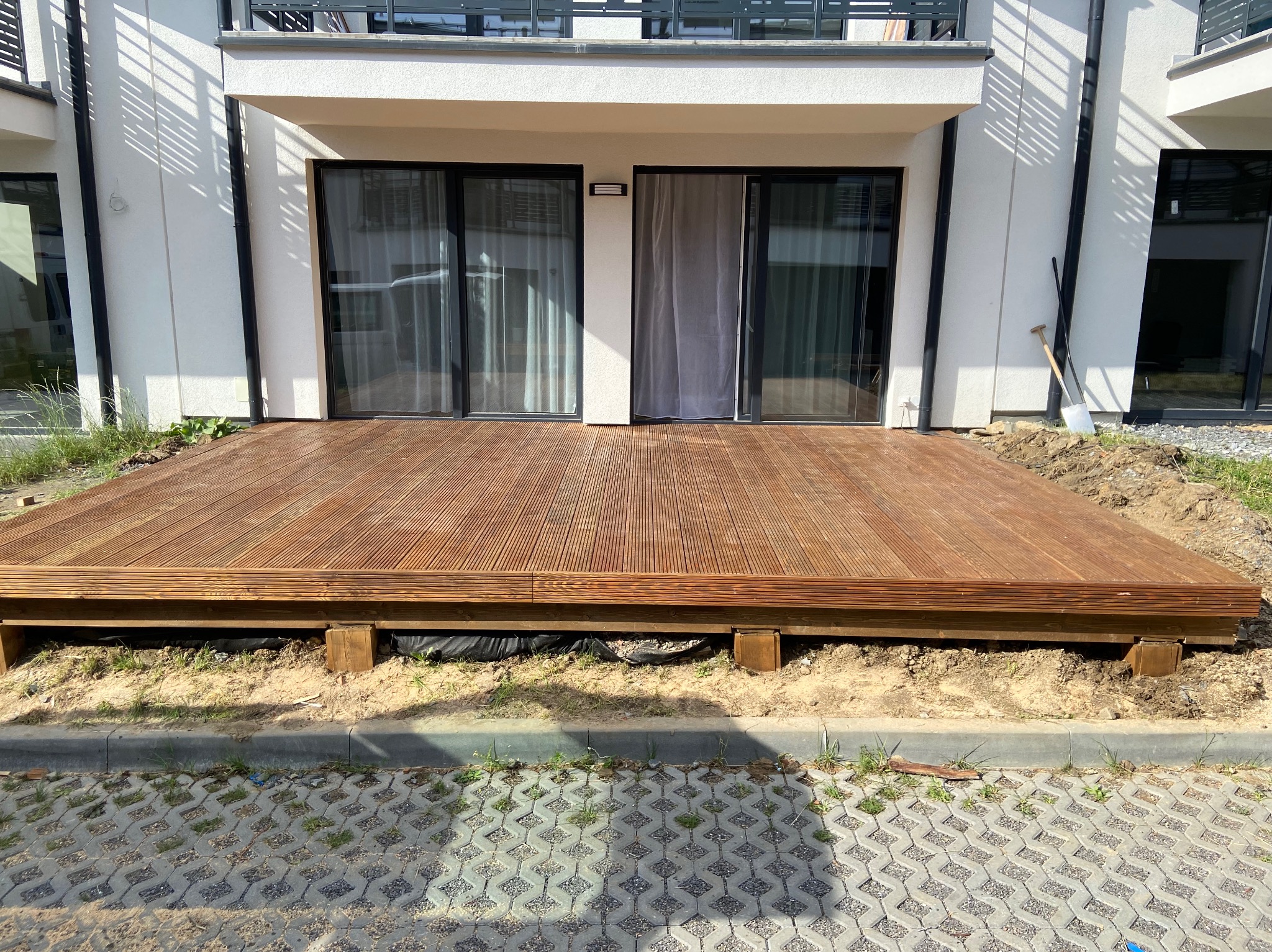 Nowy, prostokątny podest z desek na legarach, przylegający do budynku z dużymi oknami przesuwnymi, widoczna łopata i szpadel oparta o ścianę.