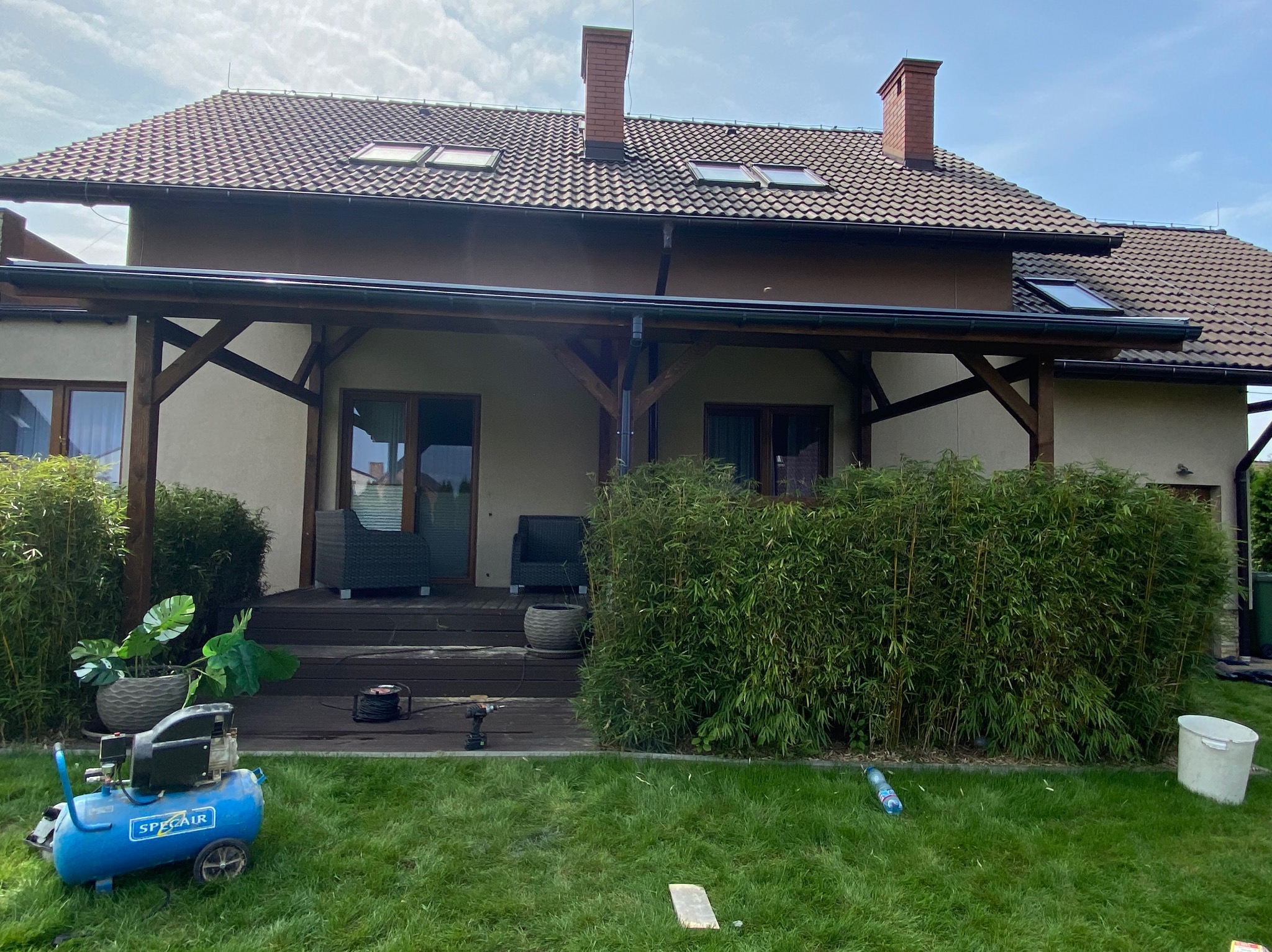 Drewniany taras z zadaszeniem przy domu, częściowo osłonięty bambusami, z widocznym kompresorem, wiertarką i doniczkami z roślinami.