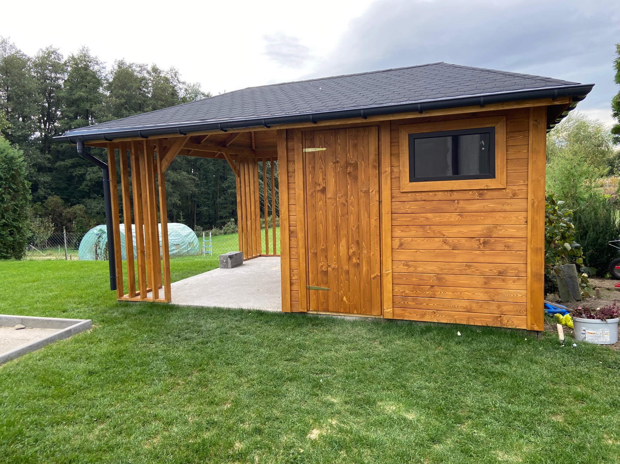 Nowoczesna, drewniana konstrukcja z zadaszeniem, przypominająca mały domek lub pawilon ogrodowy, z wyraźnymi słojami drewna, czarnym dachem i rynnami, otoczona zielonym trawnikiem i drzewami w tle.