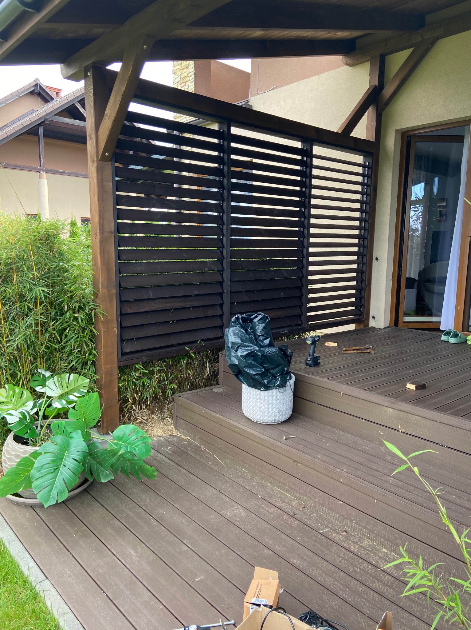 Montaż drewnianej pergoli z lamelami na tarasie: widoczne narzędzia, resztki materiałów, doniczki z roślinami, częściowo ukończona konstrukcja z ciemnego drewna.