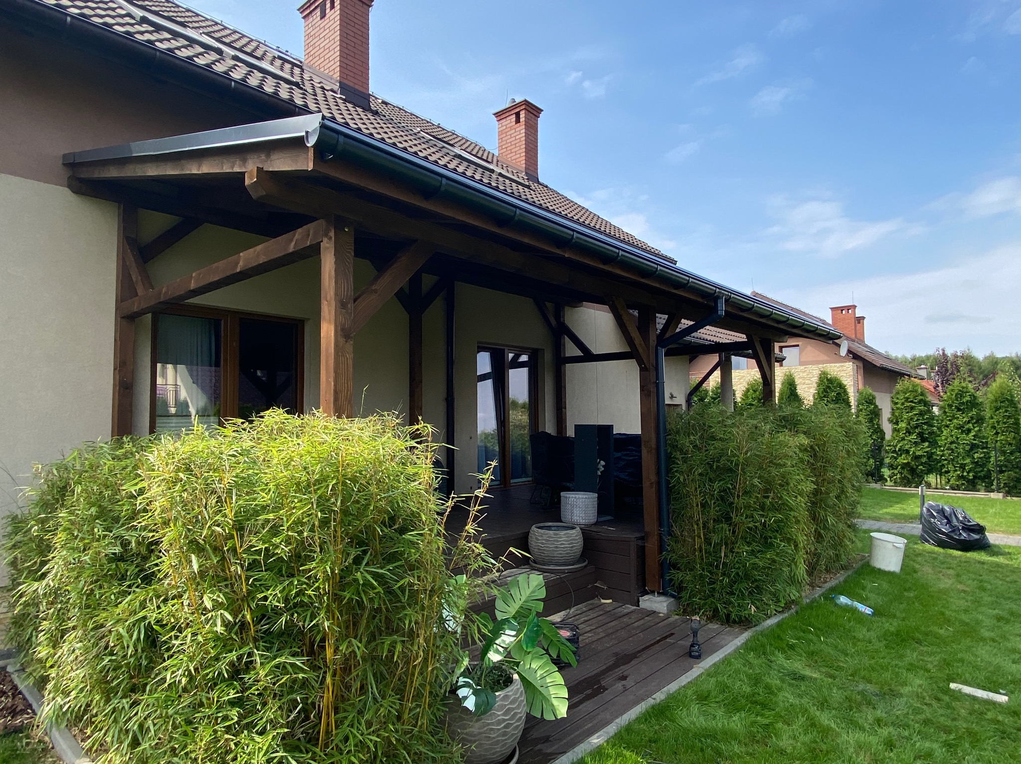 Zadaszony taras z ciemnego drewna, otoczony bambusową zielenią, z donicami i widokiem na zadbany trawnik oraz błękitne niebo.