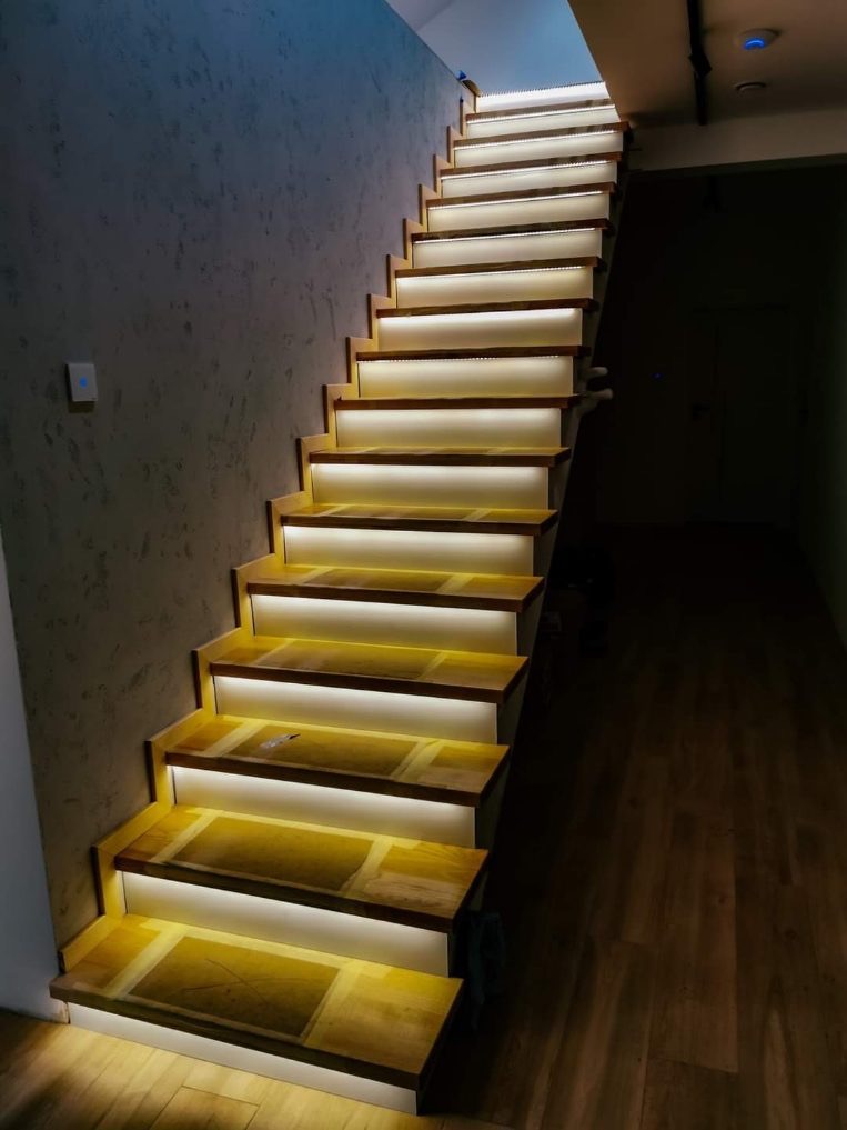 Nowoczesne schody z podświetleniem LED na każdym stopniu, widoczne z dołu do góry, z teksturą ściany po lewej stronie i ciemną podłogą po prawej.
