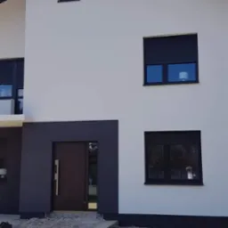 Dwukondygnacyjny dom z białymi ścianami, ciemnymi oknami z roletami zewnętrznymi i ciemnym dachem, widoczne wiadra z farbą i narzędzia malarskie przed wejściem.