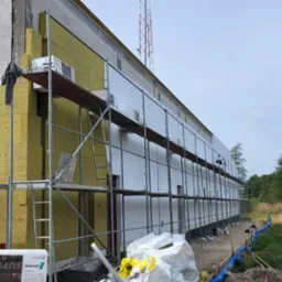 Budynek w trakcie ocieplania, widoczne rusztowania, częściowo nałożona żółta wełna mineralna i białe płyty izolacyjne, z boku paleta z materiałami izolacyjnymi.