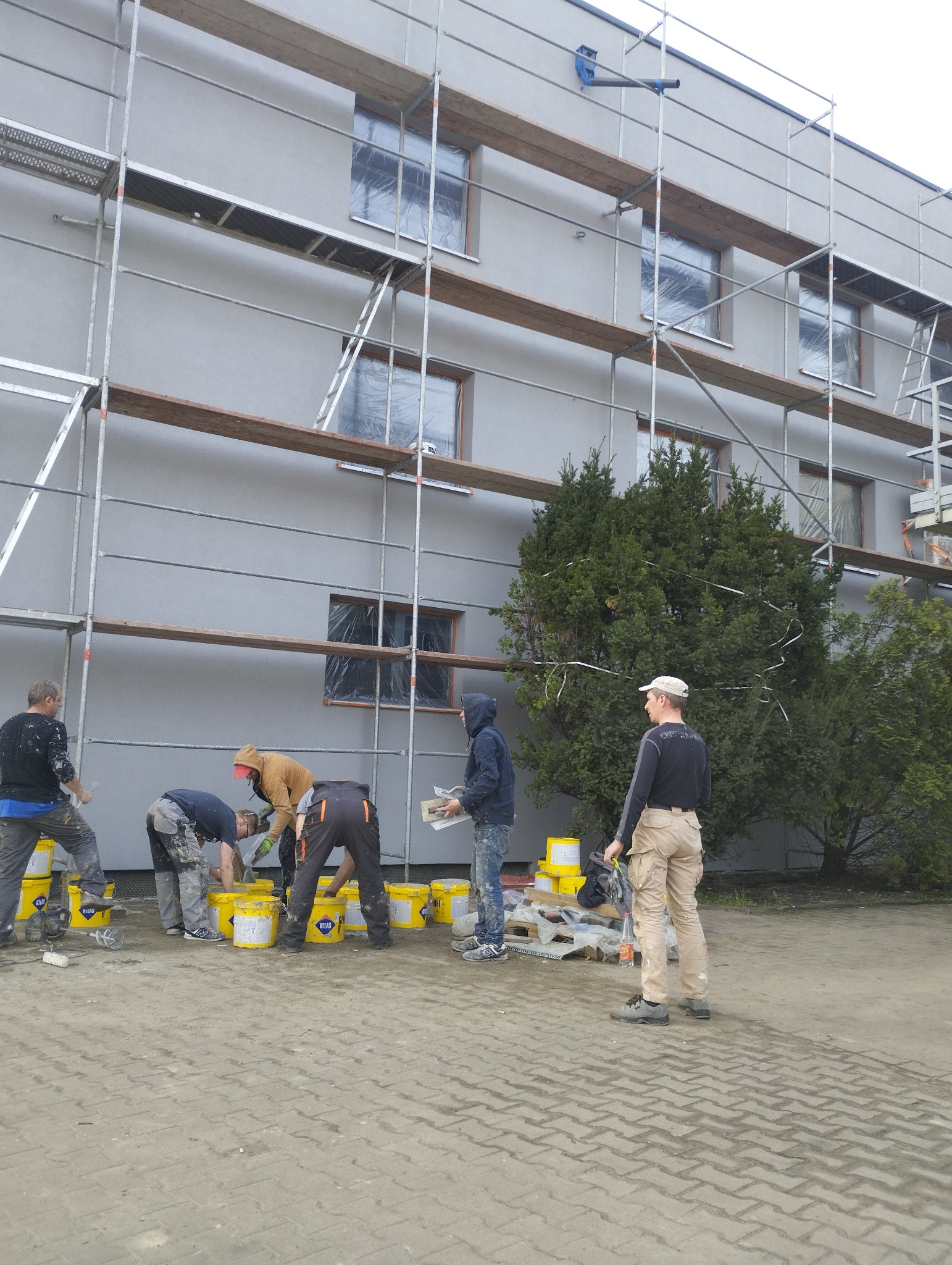 Ekipa pracowników w trakcie nakładania szarej warstwy elewacyjnej na budynek z rusztowaniem, widoczne żółte wiadra z materiałem firmy Atlas.