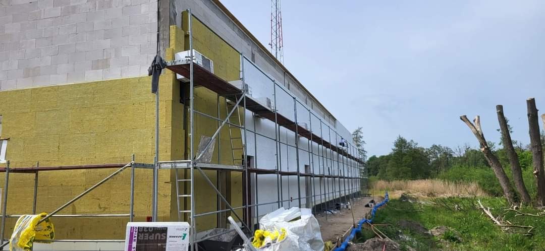 Budynek w trakcie ocieplania, widoczne rusztowania, częściowo nałożona żółta wełna mineralna i białe płyty izolacyjne, z boku paleta z materiałami izolacyjnymi.