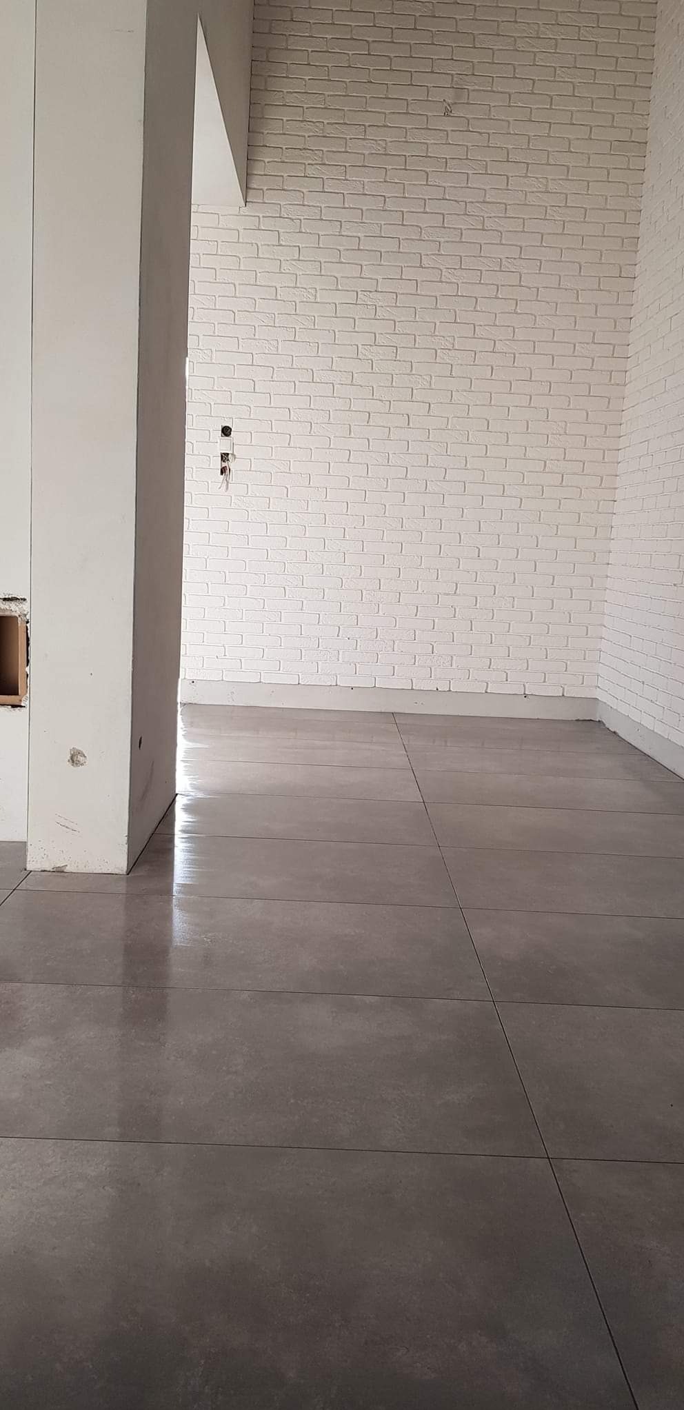 Ułożona szara podłoga z płytek w pomieszczeniu z białą cegłą na ścianie, widoczne niedokończone prace przy kolumnie i instalacji elektrycznej.